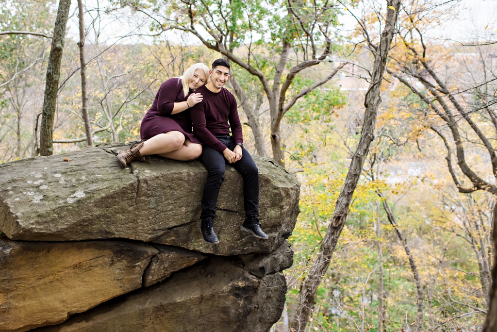 A young couple sit on a large bolder in the woods during their engagement session in Delaware.